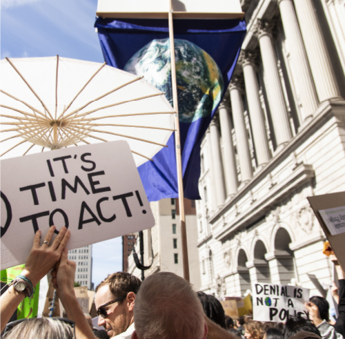 Protesters in front of a building with classical architecture, a picture of the planet on a flag, and a person holding a sign that says "It's Time to Act"